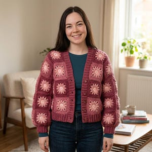 May include: A long-sleeved crochet cardigan featuring a patchwork design of square motifs. Each square displays a central light pink flower pattern surrounded by dark pink yarn. The garment has a V-neckline, a button-down front, and ribbed cuffs.