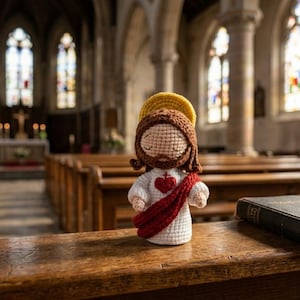 May include: A crocheted doll of a figure with brown hair, a yellow halo, and a white robe featuring a red sash and a red heart emblem, positioned on a wooden pew next to a closed black book and a string of brown prayer beads.