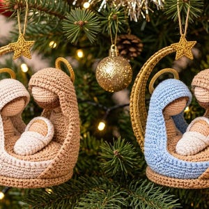 Puede incluir: Dos adornos de belén de ganchillo con figuras en hilo color canela, blanco y azul claro. Cada adorno incluye una estructura en forma de media luna rematada con una estrella dorada y una pequeña figura infantil envuelta en hilo blanco.