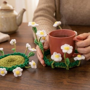 Puede incluir: Dos posavasos redondos de ganchillo verde con flores blancas y amarillas similares a margaritas en tallos. Un posavasos está plano, mientras que el otro sostiene una taza de cerámica color terracota, con las flores rodeando la base.