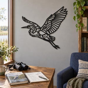 May include: A black metal wall art piece shaped like a flying heron with an intricate cutout design. The bird is depicted in mid-flight with wings spread wide, showing detailed feather patterns and a long neck, mounted on a plain wall surface.