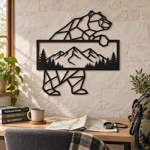 Op de afbeelding: Een zwart metalen wanddecoratie met het geometrische silhouet van een beer. In het midden van de beer bevindt zich een rechthoekig kader met een landschap van bergtoppen en een dicht dennenbos, uitgevoerd in een strakke, lineaire stijl.