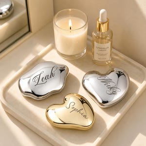 May include: Three metallic compact mirrors in heart and organic shapes, engraved with the names Leah, Nicky, and Sophia. Two mirrors are silver-toned and one is gold-toned. They are arranged alongside a lit candle and a bottle of Centella Ampoule.