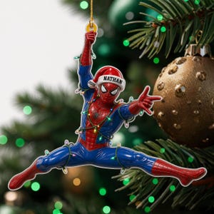 May include: A flat hanging ornament shaped like Spider-Man in a red and blue suit, wearing a red Santa hat with the name NATHAN and the year 2026 printed on it. The figure is wrapped in a string of small, colourful Christmas lights.