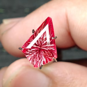 May include: A kite-shaped, vibrant pink gemstone featuring a complex, star-like precision cut with multiple facets. The stone is held in a silver-toned metal prong setting, showcasing a sharp, geometric silhouette and a brilliant, reflective surface.
