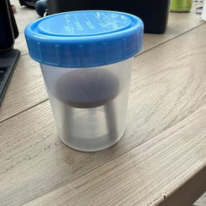 May include: A translucent plastic specimen container with a bright blue screw-on lid. The lid features printed text fields for Name, Room No, Doctor, and Date. Inside the cup, a dark circular insert is visible, suspended within the clear vessel.