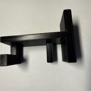 May include: A black 3D printed wall mount hook featuring a flat rectangular back plate, a horizontal shelf extending outward, and two vertical prongs extending downward from the shelf to provide support for hanging items.