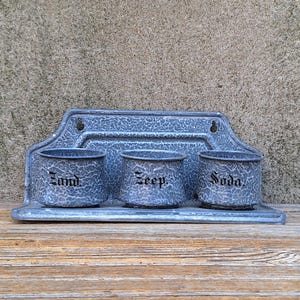 May include: A vintage, blue and gray metal wall-mounted organizer with three small, round containers. Each container is labeled with a black, gothic-style font: "Sand", "Zeep", and "Soda". The organizer sits on a wooden surface.
