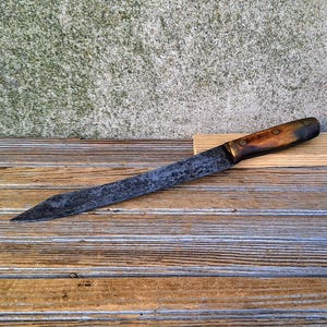May include: A long, rustic knife with a dark, weathered blade and a wooden handle. The knife rests on a small, light-colored wooden block, set against a backdrop of weathered wood and a textured wall.