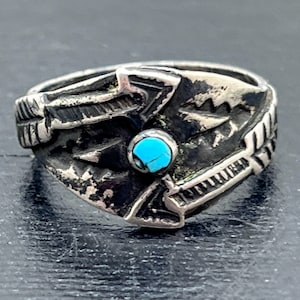 May include: A silver ring with an intricate design featuring a central turquoise stone. The ring has a detailed, textured pattern with arrow and feather motifs. The ring appears to be vintage or antique, with signs of wear.