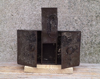 Könnte beinhalten: Eine antike Metallbox mit zwei Türen, die sich öffnen, und einem kleinen Fach in der Mitte. Die Box ist aus dunklem Metall und hat einen rustikalen Look. Sie steht auf einer Holzoberfläche.