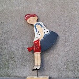 May include: A wooden figurine of a person wearing a red and white hat, a white and blue polka dot shirt, red shorts, and blue pants. The figurine is standing on a wooden base.
