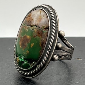 May include: A silver ring with a green and brown oval turquoise stone set in a rope-like border. The band is silver with a textured design.