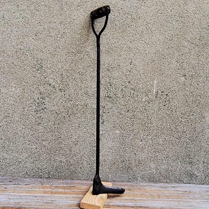 May include: A black metal fireplace poker with a D-shaped handle at the top and a pointed tip at the base. The tool stands upright, supported by a small, light-colored rectangular wooden block at its bottom, showcasing a simple, functional design.
