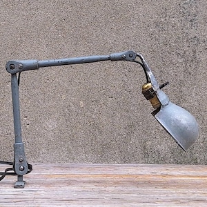 May include: A gray, adjustable desk lamp with a metal shade and a black cord. The lamp is positioned on a wooden surface against a textured gray wall. The lamp's arm is articulated for flexible positioning.