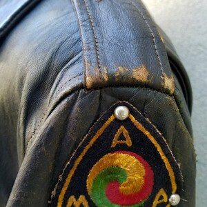 May include: A black leather jacket with a triangular patch on the left sleeve. The patch is embroidered with a gold, red and green symbol and the letters "AMA".