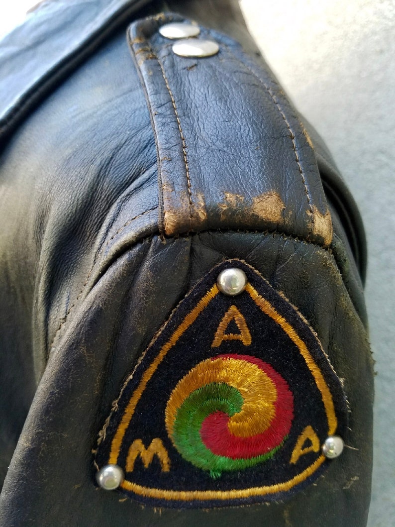 May include: A black leather jacket with a triangular patch on the left sleeve. The patch is embroidered with a gold, red and green symbol and the letters "AMA".