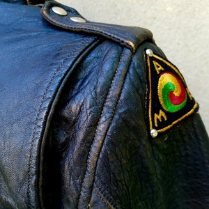 May include: Black leather jacket with a triangular patch on the shoulder. The patch is embroidered with a circle design in red, green, and yellow, and the letters "A.M." in gold thread.