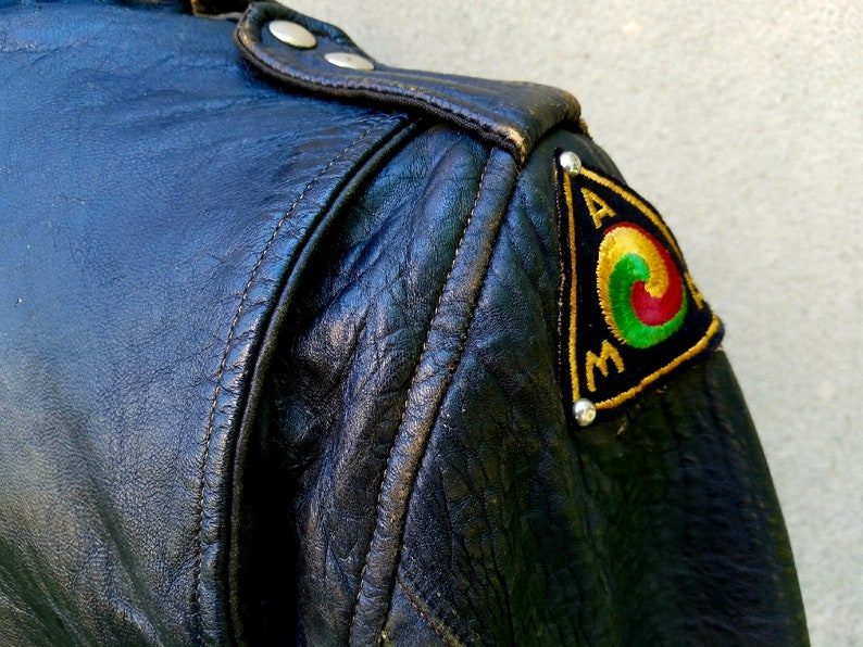 May include: Black leather jacket with a triangular patch on the shoulder. The patch is embroidered with a circle design in red, green, and yellow, and the letters "A.M." in gold thread.