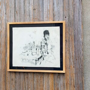 以下が含まれることがあります： テーブルにボトルとグラスを置いて座っている人の白黒の描画が額装されています。描画は白い背景に描かれており、シンプルな茶色の額縁で額装されています。