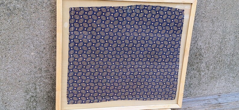 May include: A framed piece of fabric with a repeating pattern of small, light blue and gold leaves on a dark blue background.