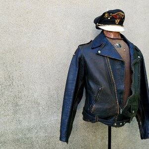 May include: A black leather biker jacket with a zip fastening and a belt at the waist. The jacket has a distressed look and is displayed on a mannequin wearing a cap.
