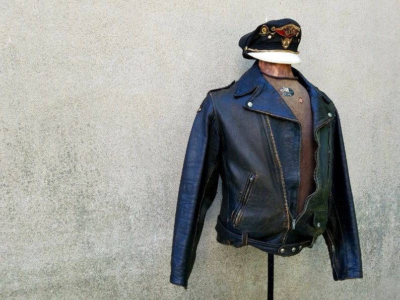 May include: A black leather biker jacket with a zip fastening and a belt at the waist. The jacket has a distressed look and is displayed on a mannequin wearing a cap.