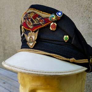 May include: A black and white cap with a white brim and a gold cord. The cap has several patches and pins, including a winged shield with a swirling design, a red, white and blue patch with a motorcycle, and a green, red and yellow pin with the number 7.