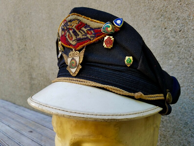 May include: A black and white cap with a white brim and a gold cord. The cap has several patches and pins, including a winged shield with a swirling design, a red, white and blue patch with a motorcycle, and a green, red and yellow pin with the number 7.