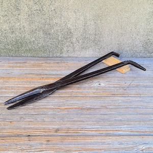 May include: A pair of antique, dark metal shears, likely for metalworking or similar tasks, resting on a weathered wooden surface. The shears have long, slender handles and a pointed cutting edge. The background is a textured, neutral-toned wall.