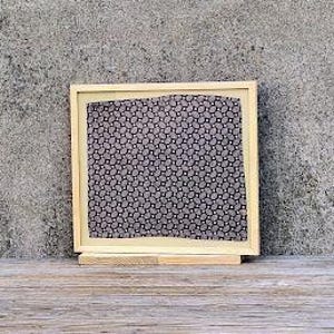 May include: A framed piece of art with a black and white geometric pattern. The frame is made of wood and has a light brown finish.