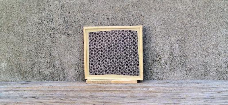 May include: A framed piece of art with a black and white geometric pattern. The frame is made of wood and has a light brown finish.