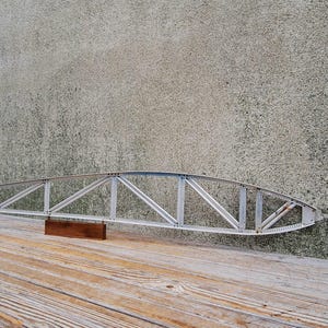 May include: A silver metal wing structure with a series of diagonal supports, resting on a small, rectangular wooden block. The wing is set against a weathered, light gray concrete wall and a wooden plank surface.