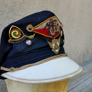 May include: A navy blue cap with a white brim and gold trim. The cap has several pins and patches, including a red, white and blue patch with the text "1962 Tour Award".