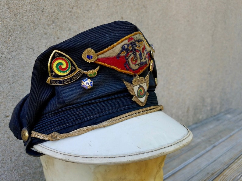 May include: A navy blue cap with a white brim and gold trim. The cap has several pins and patches, including a red, white and blue patch with the text "1962 Tour Award".