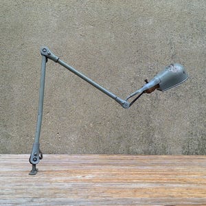 May include: A gray articulated desk lamp with a clamp base, featuring a long adjustable metal arm and a small, rounded lamp head. The lamp has visible joints and screws for positioning, finished in a matte gray tone throughout the entire structure.