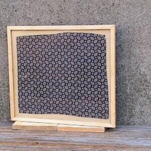 May include: A framed piece of art with a black and white geometric pattern. The frame is made of wood and has a light brown finish. The art is displayed on a wooden stand.