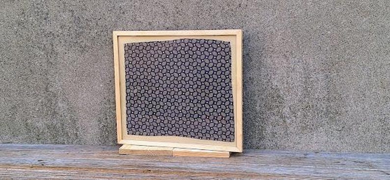 May include: A framed piece of art with a black and white geometric pattern. The frame is made of wood and has a light brown finish. The art is displayed on a wooden stand.
