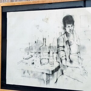 以下が含まれることがあります： テーブルにボトルやその他の物体を置いて座っている人の白黒のチャコール画。絵は木製の額縁に入っています。