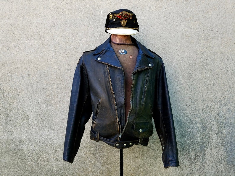 May include: A brown leather motorcycle jacket with a zip fastening and a belt at the waist. The jacket has a distressed look and is worn on a mannequin wearing a black and white cap.