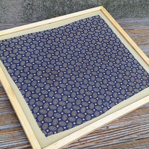 May include: A framed fabric with a blue and gold paisley pattern. The fabric is displayed on a wooden surface.