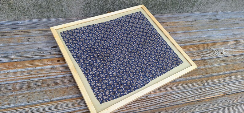 May include: A framed fabric with a blue and gold paisley pattern. The fabric is displayed on a wooden surface.