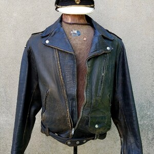 May include: A dark brown leather motorcycle jacket with a zip closure, a belt at the waist, and a zipped pocket on the left side. The jacket has a worn look and is displayed on a mannequin wearing a brown sweater and a black and white cap with gold accents.