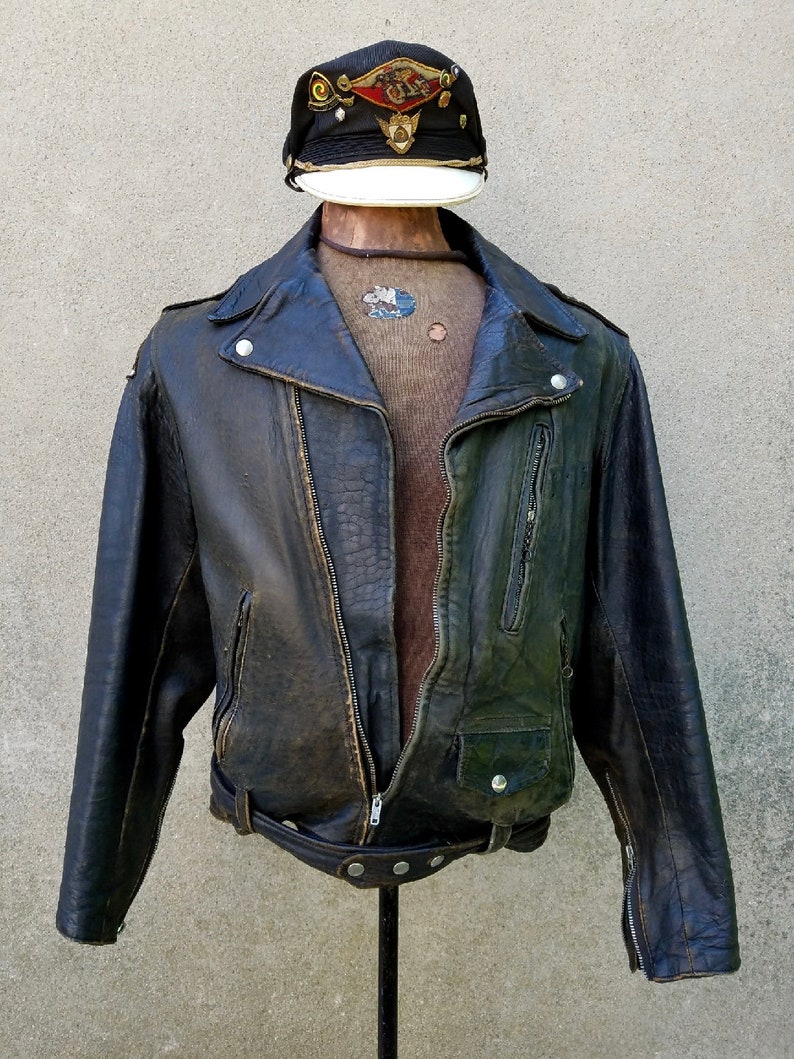 May include: A dark brown leather motorcycle jacket with a zip closure, a belt at the waist, and a zipped pocket on the left side. The jacket has a worn look and is displayed on a mannequin wearing a brown sweater and a black and white cap with gold accents.