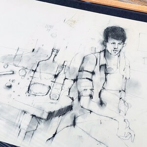 以下が含まれることがあります： テーブルにボトルを置いたテーブルに座っている人の白黒木炭画が額装されています。絵は白い紙に描かれており、明るい木製の額縁に入っています。