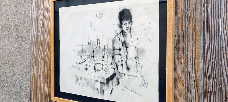 以下が含まれることがあります： テーブルにボトルとグラスを置いて座っている人の白黒の絵。絵は木製の額縁に入っています。