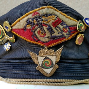 May include: A dark blue cap with a variety of pins and badges. The badges include a motorcycle design, a winged shield with a spiral design, and a small badge with the number 3. The cap has a gold braid around the brim.