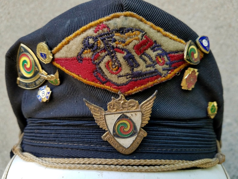 May include: A dark blue cap with a variety of pins and badges. The badges include a motorcycle design, a winged shield with a spiral design, and a small badge with the number 3. The cap has a gold braid around the brim.