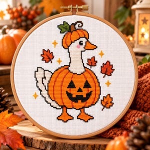 May include: A cross-stitch embroidery in a round wooden hoop featuring a white goose wearing a carved orange pumpkin as a body and a smaller pumpkin as a hat. The design includes orange autumn leaves and small yellow stars on a white fabric background.