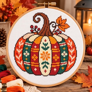 May include: A circular wooden embroidery hoop containing a cross-stitch pattern of a pumpkin. The pumpkin features vertical panels in orange, red, and teal, decorated with white and yellow floral and geometric motifs, topped with a brown stem.